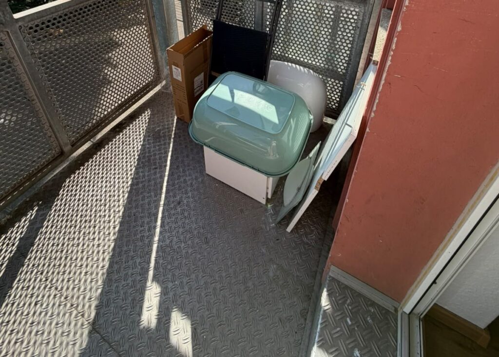 Ecke meines hinteren Balkons vor dem Umbau: Kartons, eine Kiste und ein Brett am Geländer aus Lochblech
