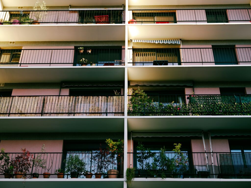 Hausfassade mit Balkonen, an den Geländern Pflanzen, eine Schilfmatte und ein grünes Rankgitter